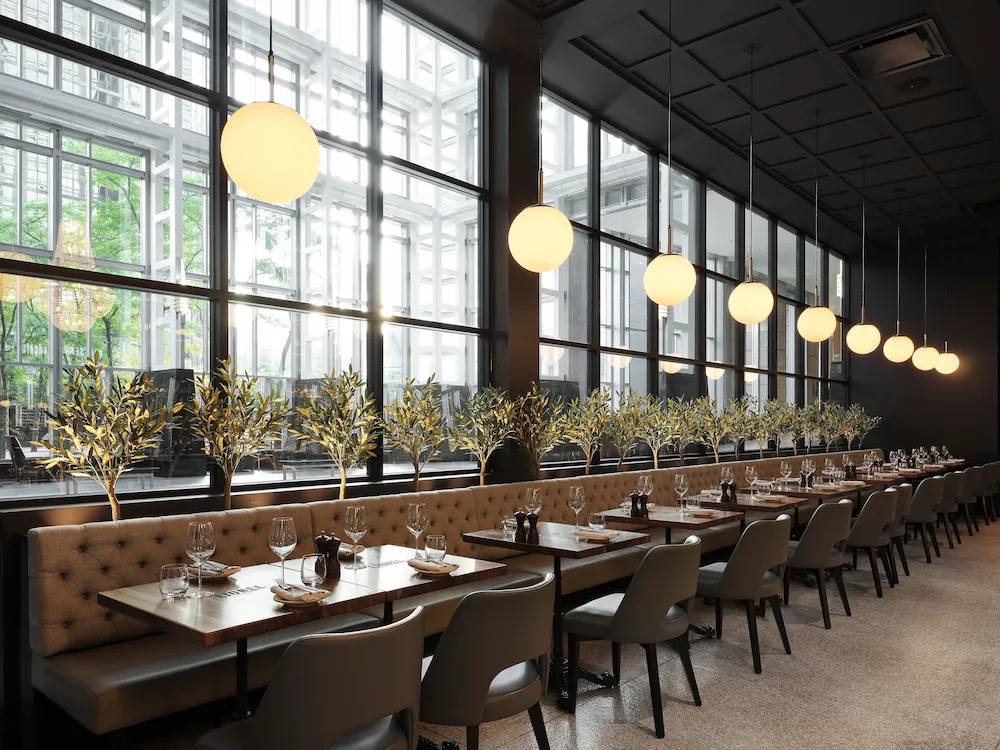 Restaurant Dining Area with a Long Row of Tables Beside Large Windows, Tufted Beige Benches, Gray Chairs, and Warm Globe Pendant Lights Above.