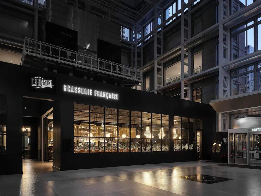 Lionel Brasserie Française Storefront with Illuminated Signage and a Warmly Lit Dining Room Visible Through Glass Walls.