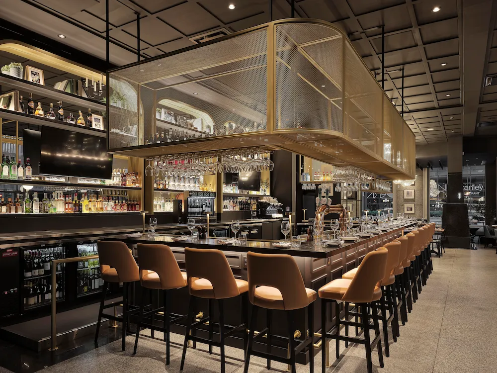 Modern Bar with a Long Black Counter, Tan Stools, and a Suspended Glassware Rack Above.