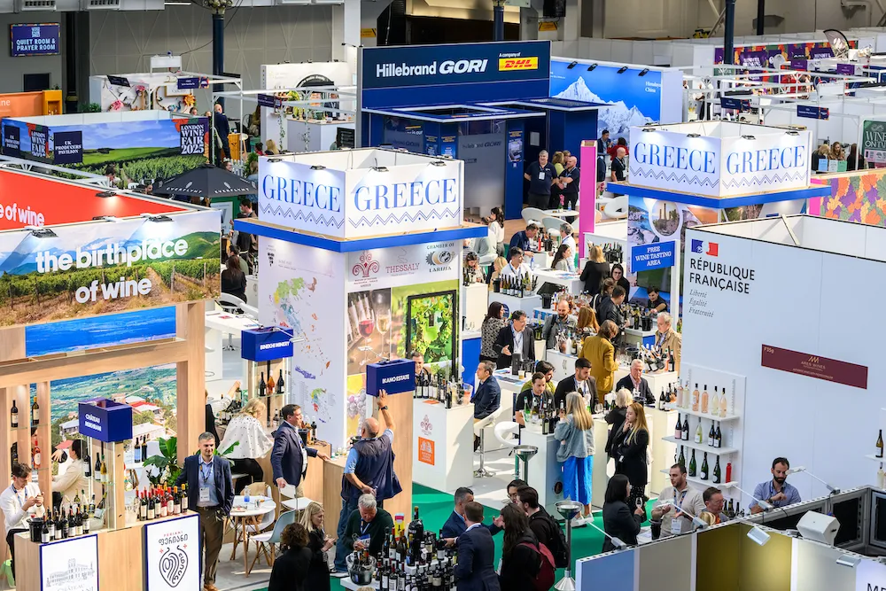 Busy Trade Fair Hall with Greece Wine Booths, Shelves of Bottles, and Visitors Mingling Around Exhibits on Display.