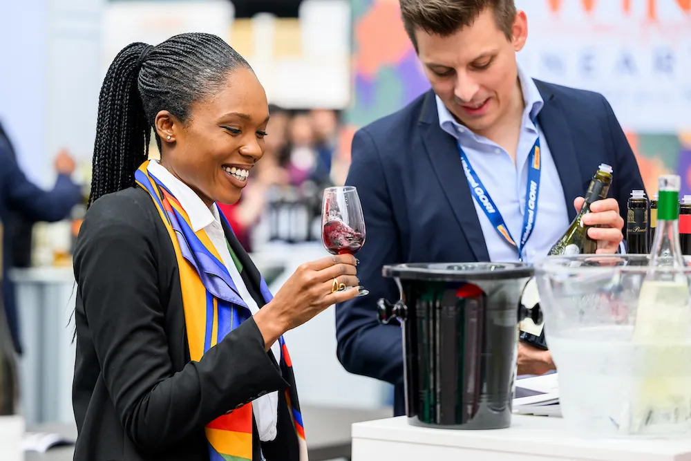 Smiling Woman in a Blazer Holding a Glass of Red Wine at a Tasting Event, with a Man Pouring Bottles Nearby.